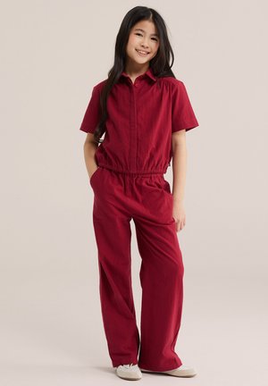 Noor tüdruk naeratab, seisab käed taskutes, seljas punane lühikeste varrukatega kombinesoon ja jalas valged jalanõud, taust neutraalne.