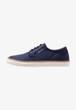 Zapatilla deportiva azul marino de caña baja con suela de goma blanca, cordones delanteros, ribetes marrones y logotipo "GANT" en el lateral.