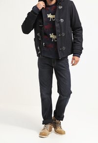 Cappotto duffle in misto lana nero con chiusure a gancio, maglione a motivi blu navy, jeans scuri e stivaletti beige con lacci.