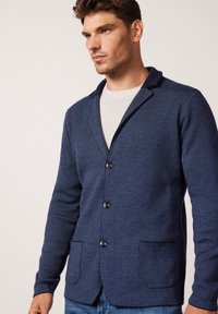 Dunkelblauer Cardigan mit Kragen, vier Knöpfen und zwei Vordertaschen; strukturierter Stoff, lässiges Design. Über einem hellen Pullover getragen.
