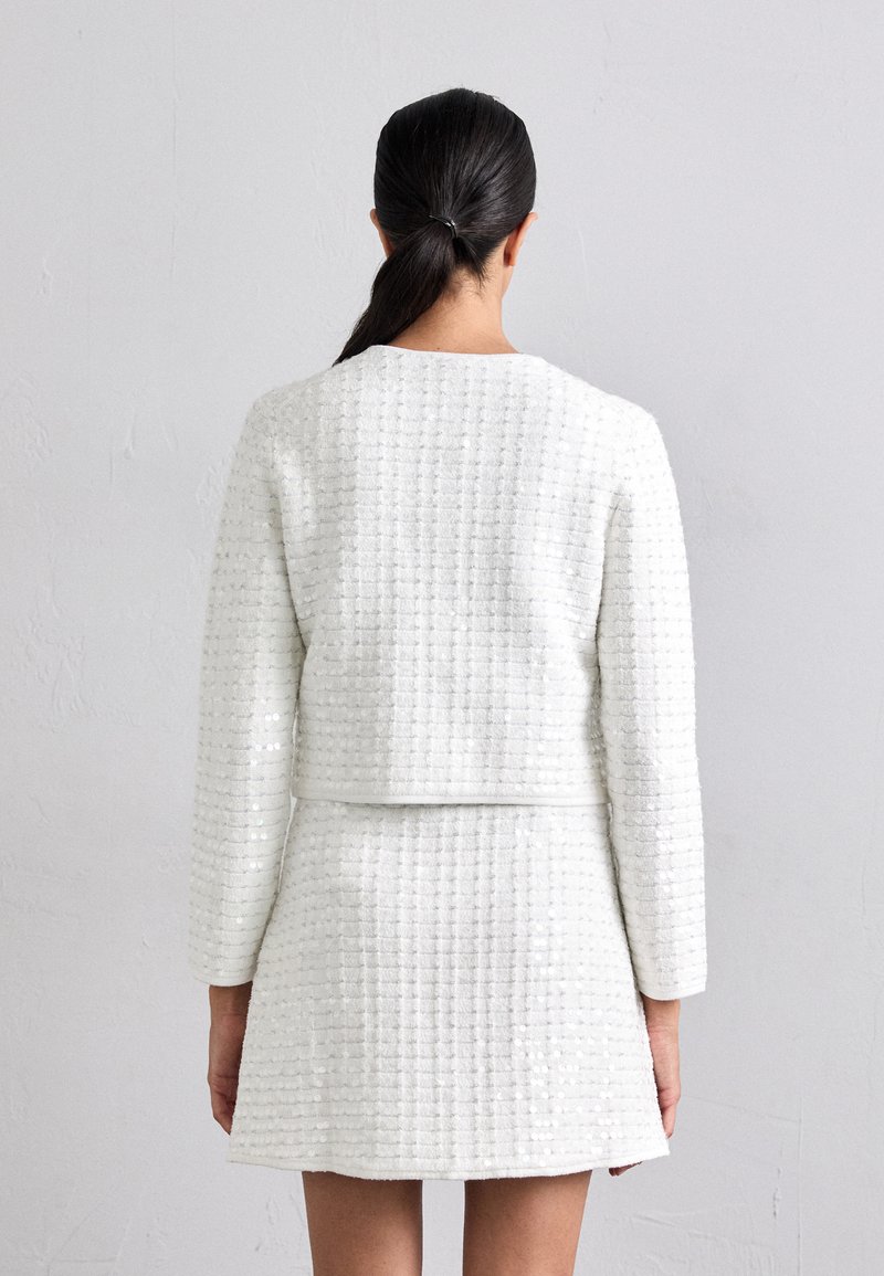 Costume deux pièces en bouclê blanc avec un motif texturé, comprenant une veste ajustée et une jupe courte, conçue avec des manches longues.