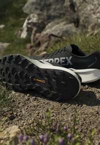 Ανδρικά παπούτσια trail running Terrex Agravic 4 σε ασπρόμαυρο χρώμα με σόλα Continental, που ακουμπούν σε χαλίκι με βράχια και γρασίδι στο φόντο.