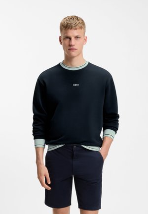 Νέος άνδρας με κοντά ξανθά μαλλιά, φοράει μαύρο φούτερ με λογότυπο "BOSS", ανοιχτοπράσινες μανσέτες και σκούρα σόρτς, στεκόμενος με το ένα χέρι στην τσέπη.