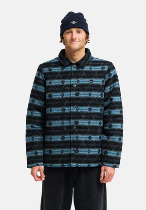 Mann, der eine gemusterte blau-schwarze Jacke mit Knöpfen und eine dunkelblaue Mütze trägt, steht und schaut mit einem leichten Lächeln nach vorne.