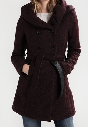Woman wearing a dark purple double-breasted wool coat with wide collar, belted waist, and hands in pockets, paired with blue jeans.