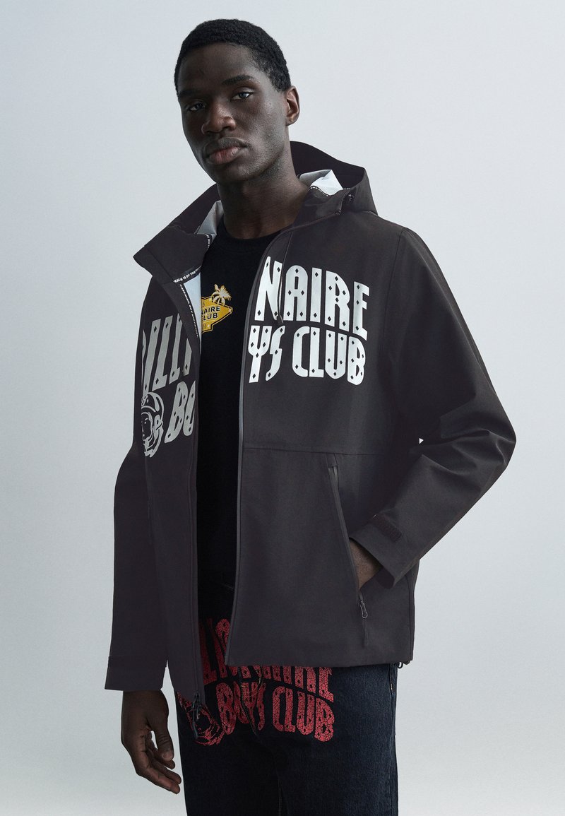 Μαύρο μπουφάν με μεγάλα λευκά γράμματα και γραφικά στοιχεία. Διαθέτει τσέπες με φερμουάρ, κουκούλα και λεία υφή ύφασματος.