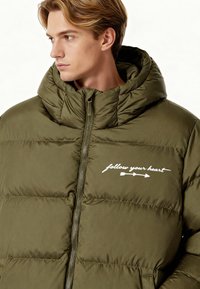 Giacca imbottita verde oliva con collo alto, dotata di una zip frontale e del testo bianco "segui il tuo cuore" lungo il petto. Texture liscia.