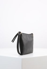 Liten svart läderhandväska med dragkedja och fäst handledsrem, uppvisad på en vit yta mot en enfärgad vit bakgrund.