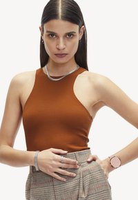 Roodbruine mouwloze top van gestructureerde stof, gedragen met een hooggetailleerde geruite rok. Zilveren sieraden en een roze horloge maken de look compleet.