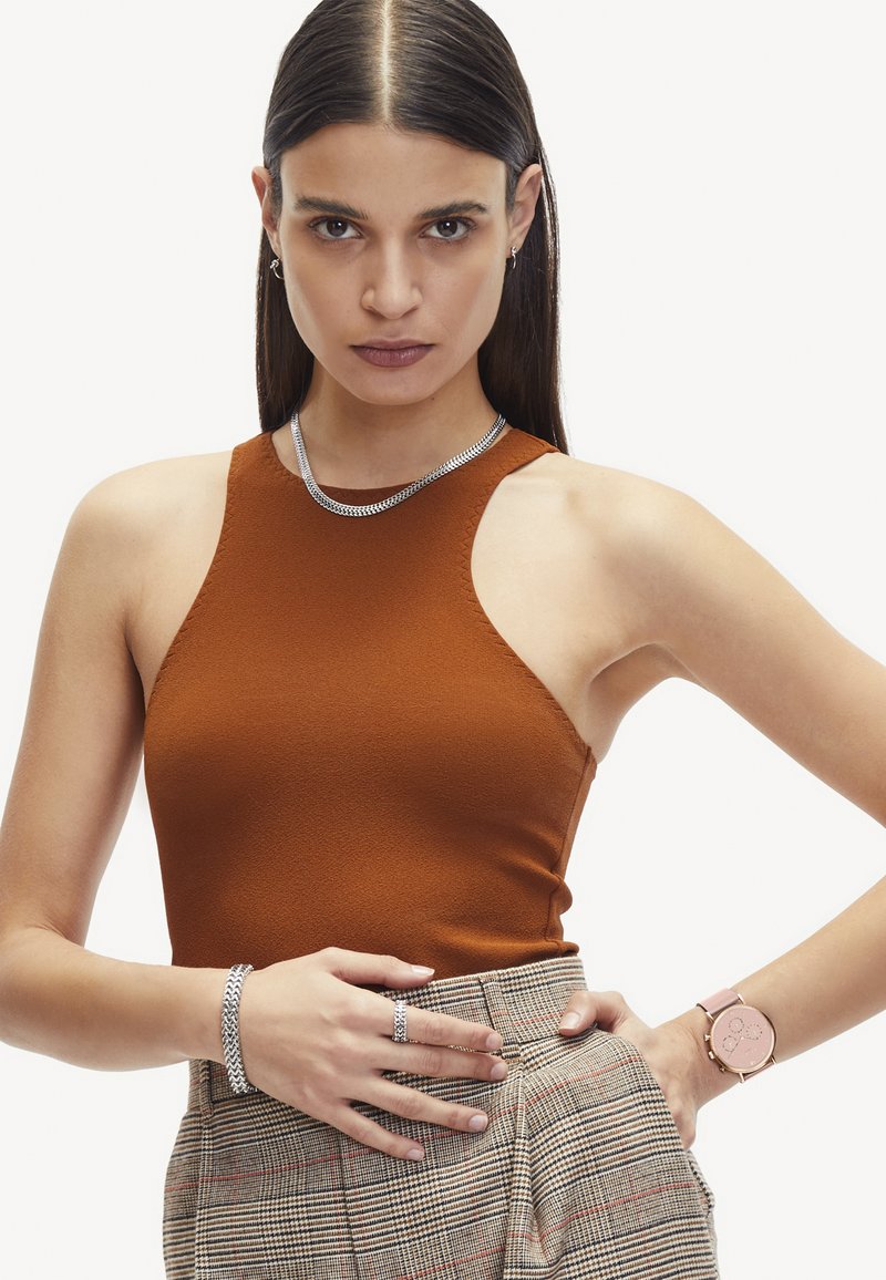 Roodbruine mouwloze top van gestructureerde stof, gedragen met een hooggetailleerde geruite rok. Zilveren sieraden en een roze horloge maken de look compleet.