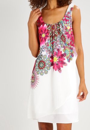 Kvinde iført hvid ærmeløs kjole med lagdelt kant og livlige pink, røde og blå blomstrede mandalamønstre foran.