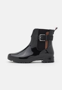 Bottes de pluie noires brillantes jusqu'à la cheville avec des panneaux élastiques sur les côtés, un détail de boucle et une semelle texturée pour l'adhérence. Présente un logo sur la sangle arrière.