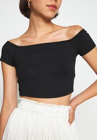 Černý crop top s odkrytými rameny vyrobený z měkké bavlny, s krátkými rukávy a fitted designem, spárovaný s bílou texturovanou sukní.