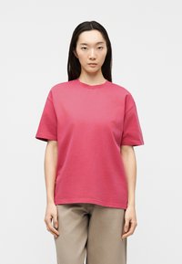 Νέα γυναίκα με μακριά σκούρα μαλλιά, φορώντας ένα απλό ροζ κοντομάνικο μπλουζάκι και μπεζ παντελόνι, στέκεται με ουδέτερη έκφραση.