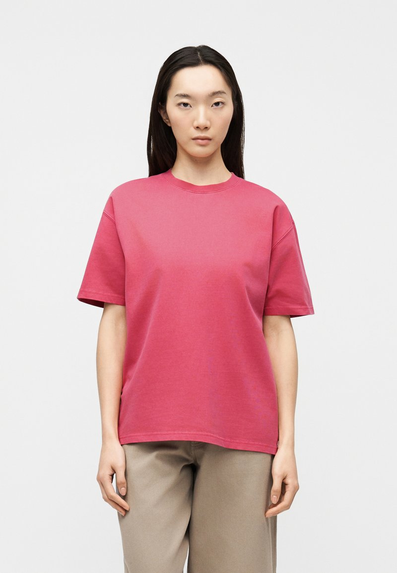 Νέα γυναίκα με μακριά σκούρα μαλλιά, φορώντας ένα απλό ροζ κοντομάνικο μπλουζάκι και μπεζ παντελόνι, στέκεται με ουδέτερη έκφραση.