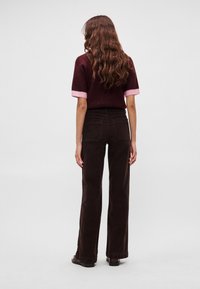 Καφέ παντελόνι από κορντόν με φαρδιά γραμμή και πίσω τσέπες· συνδυασμένο με μπορντό μπλούζα που έχει ροζ μανσέτες στα μανίκια. Μακριά κυματιστά μαλλιά εμφανή.