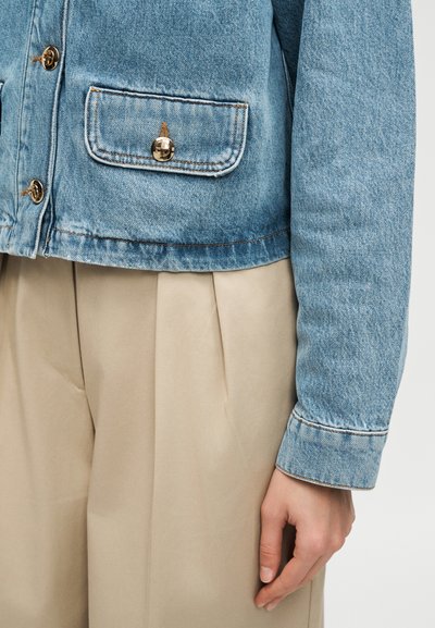 Ανοιχτό μπλε τζιν μπουφάν με ορειχάλκινα κουμπιά πάνω από μπεζ πλισέ παντελόνια, εμφανίζοντας το δεξί χέρι και το χέρι του ατόμου χαλαρά στο πλάι.