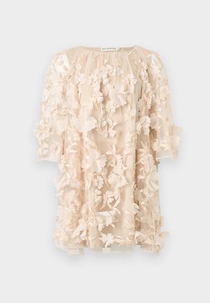 Femme portant une robe beige transparente mi-longue ornée d'appliqués floraux en 3D et des sandales beiges à talons, debout sur un fond blanc uni.