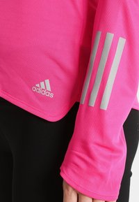 Zbliżenie na osobę w jasnoróżowej bluzie Adidas z długim rękawem, z odblaskowymi trzema pasek na rękawie oraz czarnymi spodniami.