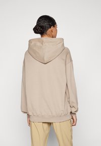 Beige hoodie met een grote, gevoerde capuchon, oversized pasvorm en geribde manchetten en zoom. Vloeibare textuur tegen een effen achtergrond.