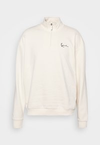 Sudadera color crema con un cuello alto, cremallera en la parte frontal hasta la mitad, mangas largas, puños acanalados y un simple logotipo negro en el pecho.