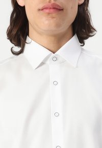 Chemise blanche avec un tissu texturé, un col pointu et des boutons accentués de bleu. La coupe semble ajustée, avec une finition lisse.