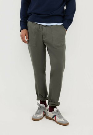 Hombre con pantalones verde oliva remangados, zapatillas blancas con rayas rojas y azules y calcetines rojo oscuro, de pie contra un fondo blanco.