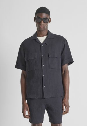 Man draagt zwart gestructureerd kortärmelig overhemd met knopen en twee borstzakken, bijpassende korte broek en donkere oversized zonnebril tegen een grijze achtergrond.