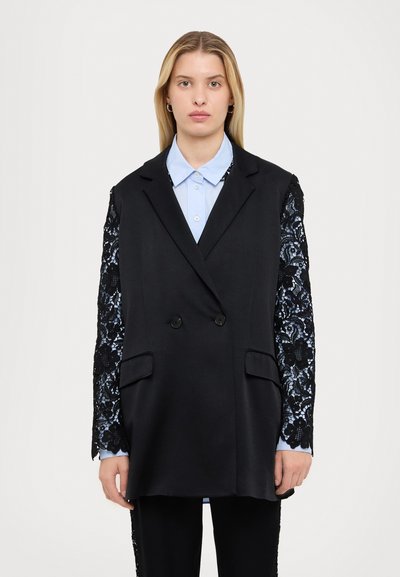 Black tailored blazer with lace sleeves, double-breasted design, two front pockets, worn over a light blue shirt. Textured floral lace detail.