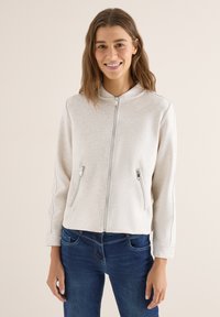 Femme aux cheveux bruns mi-longs portant une veste beige clair zippée avec des poches zippées et un jean bleu foncé, souriante, debout devant un fond uni.