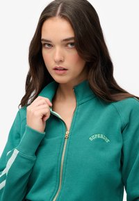 Sweat-shirt zippé bleu canard avec fermeture éclair dorée, poignets côtelés et logo brodé "SUPERDRY". Tissu doux, design à col haut, rayures latérales subtiles.