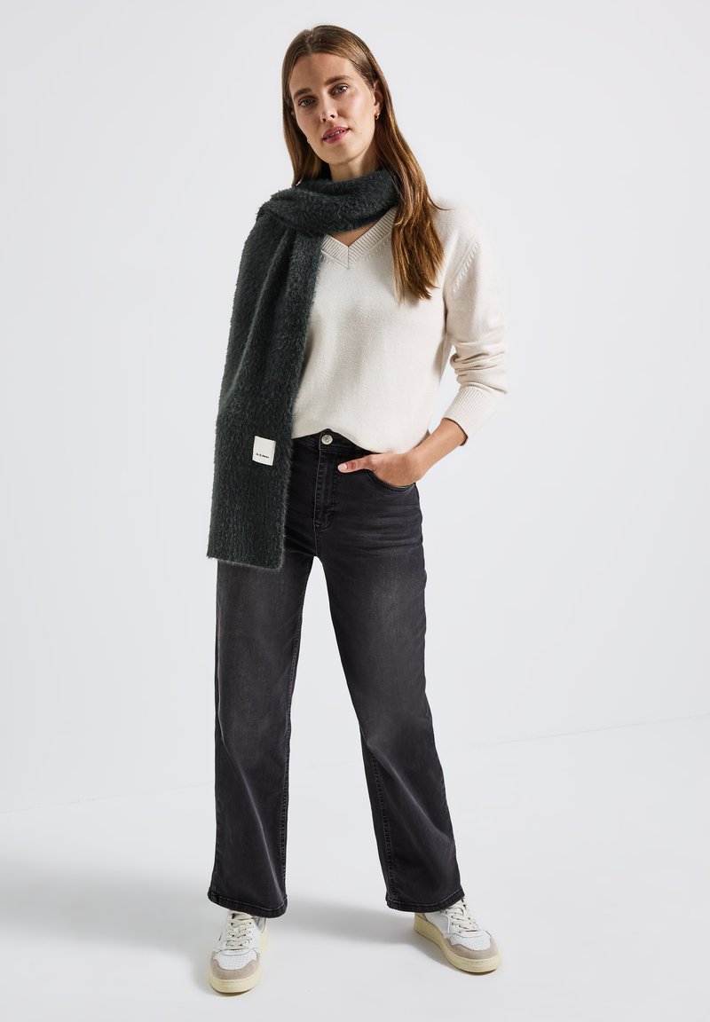 Bufanda de piel sintética verde oscuro, suéter de punto color crema y pantalones de mezclilla anchos negros. Zapatillas con suela texturizada y acentos sutiles. Diseño simple.