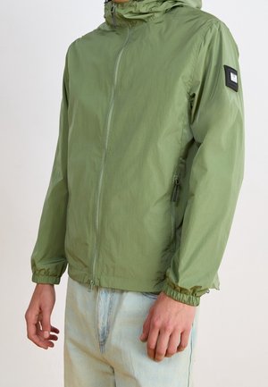 Osoba ubraną w jasną zieloną kurtkę przeciwwiatrową z kapturem na zamek błyskawiczny, z elastycznymi mankietami i jasnoniebieskie dżinsy, stoi z rozluźnionymi ramionami.