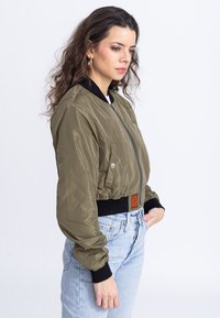 Giacca bomber verde oliva, finitura in satin, collo e polsini a coste neri, chiusura con zip frontale, tasche laterali, design cropped con dettagli arancioni.
