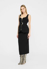 Schwarzes ärmelloses Kleid mit enganliegendem Oberteil, Reißverschluss vorne und breitem Gürtel; kombiniert mit metallischen Schlangenleder-Stiefeletten. Glattes, strukturiertes Material.