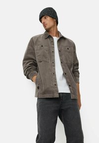 Grå kordfløjlsjakke med to brystlommer og sorte knapper, båret over en hvid skjorte. Puslet med mørke jeans og en sort strikket hue.