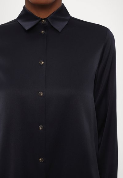 Woman wearing a black satin long-sleeve button-up shirt with a pointed collar against a plain background.