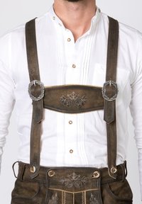 Bruna läderhosen med silverdetaljer, dekorativt stygn och justerbara remmar över en vit plisserad skjorta med dekorativa knappar.