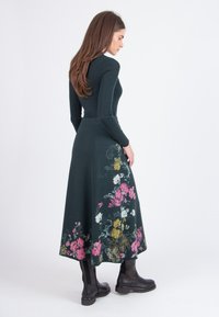 Lengte lange, donkergroene jurk met ribbelstructuur, met kleurrijke bloemenpatronen in roze, wit en geel langs de zoom. Zwarte kniehoge laarzen.