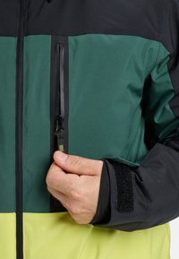 Color-blocked jacket featuring black, green, and yellow panels. Includes a zippered pocket and adjustable Velcro cuffs. Smooth texture.