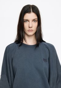 Veľký tmavomodrý sweatshirt s rebrovanou textúrou, okrúhlym výstrihom a vyšitým logom na hrudi. Má spustené ramená a dlhé rukávy.