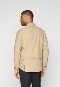 Vaalean beige kevyt takki, jossa on pystykaulus, joustavat hihansuut ja vetoketjullinen tasku selässä. Raskaampi kangas, jossa on hieno kiilto.