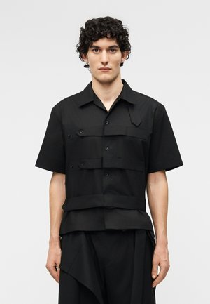 Jeune homme aux cheveux courts, bouclés et foncés, portant une chemise noire à manches courtes avec des détails de tissus superposés et un pantalon noir assorti.