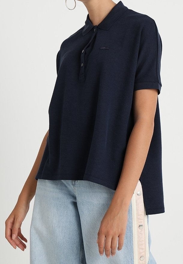 Femme portant un polo bleu marine à manches courtes et un jean bleu clair avec des détails de boutons sur le côté, debout devant un fond uni.