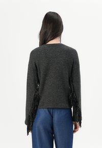 Grauer Pullover mit langen schwarzen Fransen an den Ärmeln, Rundhalsausschnitt und lässiger Passform, kombiniert mit blauen Hosen.
