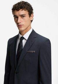 Abito blu navy con taglio slim, rever e una camicia bianca. Presenta una cravatta a righe diagonali e un fazzoletto da taschino a fantasia nella tasca del petto.