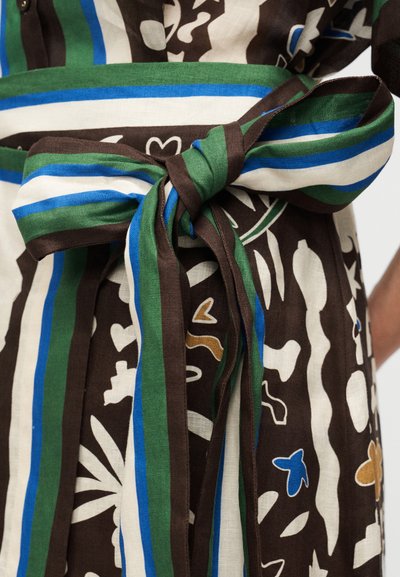 Υφασμάτινη ζώνη που δένεται στη μέση με ρίγες σε πράσινο, μπλε, λευκό και καφέ χρώμα πάνω από καφέ ένδυμα με λευκά, μπλε και μπεζ αφηρημένα σχήματα.