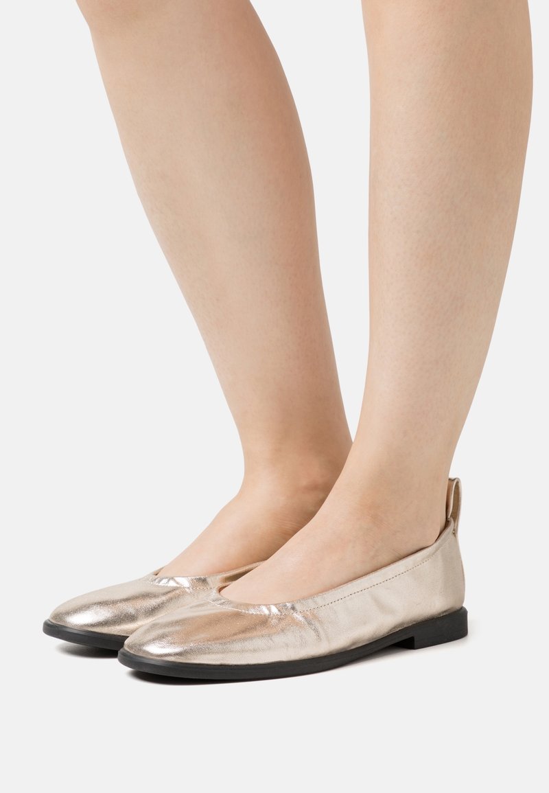 Jambes portant des ballerines dorées métalliques avec des semelles noires sur un fond clair uni.