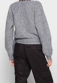 Grå stickad tröja med ett strukturerat mönster, ribbade ärmslut och en knytning i midjan, tillsammans med mörka cargo-byxor.