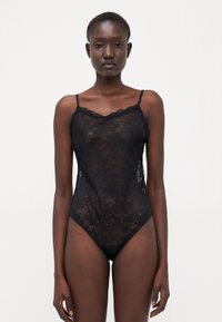 Bodysuit en dentelle noire avec de fines bretelles et des bords festonnés. Présente un motif floral et des jambes échancrées. Texture transparente et design ajusté.